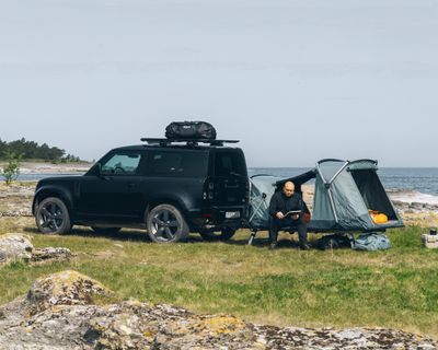 A  man sits inside his towbar tent that is attached to his vehicle parked next to the water.